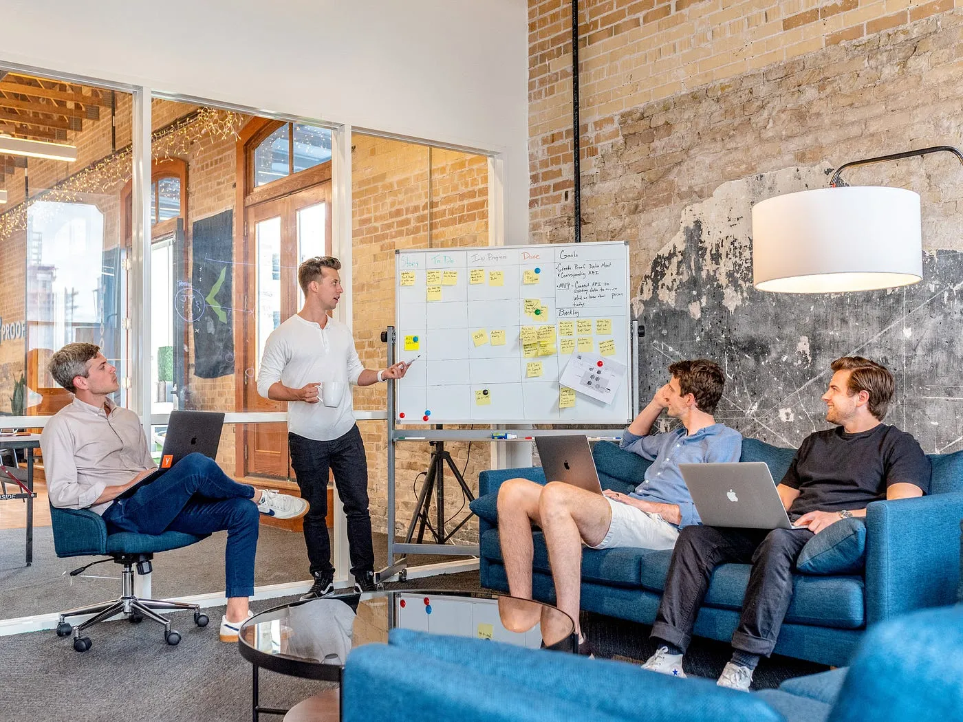 Team meeting in an office: one person presents at a whiteboard covered in sticky notes while the others listen with laptops